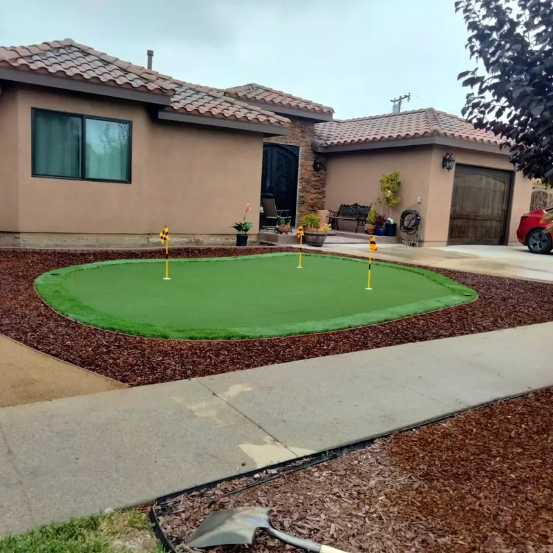 A synthetic putting green with three flag cups set into a front yard, edged in red bark mulch