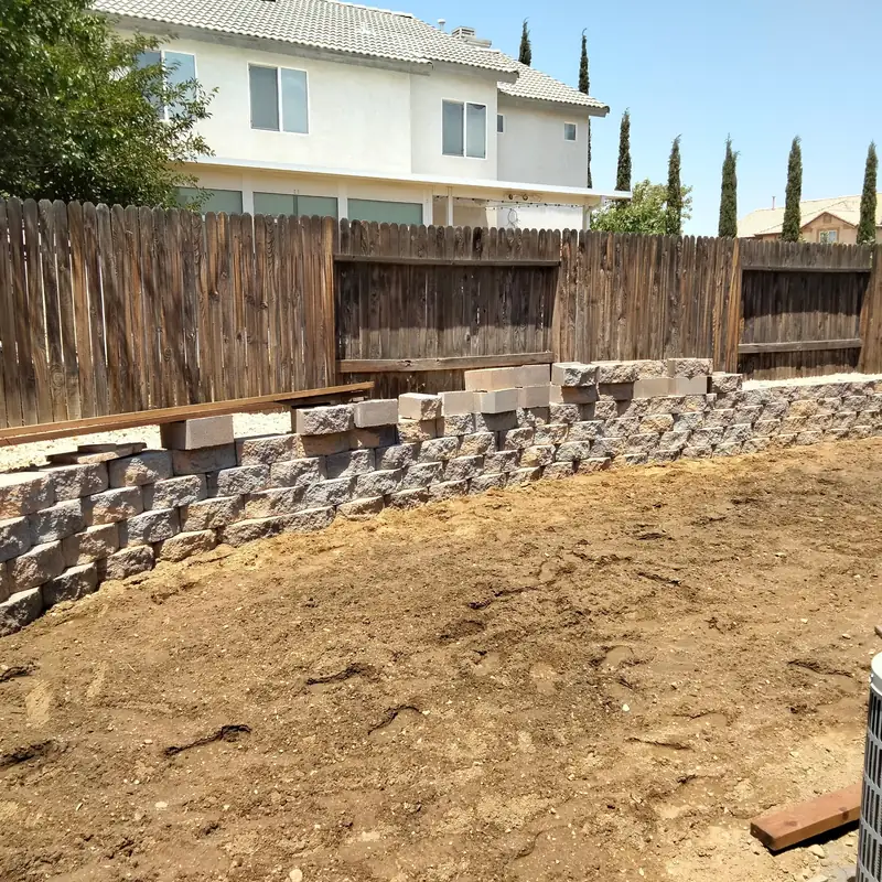 A split-face block retaining wall being built in courses along the back of a yard, with the ground behind it still open